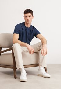 Navy blue cotton t-shirt with a small black pocket. Paired with beige chinos and white sneakers. Model sitting on a light beige chair.