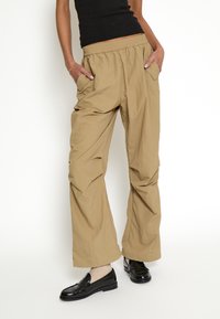 Pantaloni beige a gamba larga con vita elasticizzata, due tasche laterali, tessuto leggero, trama liscia, abbinati a mocassini neri e calzini a fantasia.