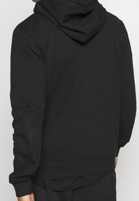 Bakvy av en person som bär en svart hoodie med långa ärmar och svarta byxor, stående mot en enkel ljus bakgrund.