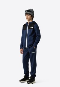 Navy tracksuit with black accents, zip-up jacket, elastic cuffs, and matching pants. White logo displayed on the chest and thigh. Gray beanie and sunglasses.