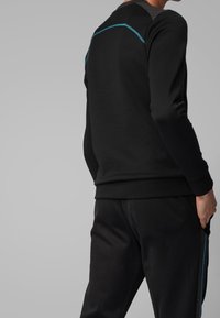 Felpa nera con scollo rotondo, polsini a costine e un accento turchese sulla schiena. Abbinata a pantaloni neri coordinati.