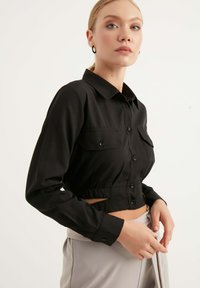 Camicia nera cropped con bottoni e colletto, due tasche sul petto e maniche lunghe. Il tessuto sembra liscio con un design aderente in vita.