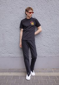 Camiseta de algodón negra con un gráfico amarillo y rojo en el pecho, combinada con jeans negros y zapatillas blancas. El modelo está de pie frente a una pared gris.