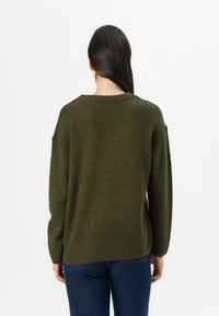 Olivenfarget oversized genser laget av myk, teksturert stoff, med vid halsutskjæring og ribbestrikket kant, sammen med mørkeblå jeans.