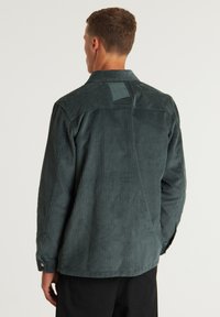 Donkergroene corduroy overhemd met verticale ribbels, knoopmanchetten en een yoke detail aan de achterkant. Bevat een kleine geborduurde patch op de kraag.