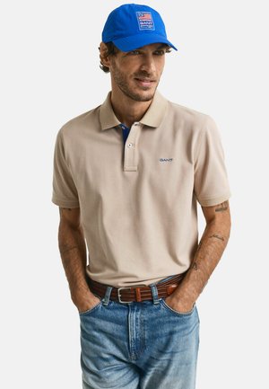 Polo beige avec un col bleu et le logo GANT. Associé à un jean en denim bleu et une casquette bleue avec un patch logo. Ceinture marron.
