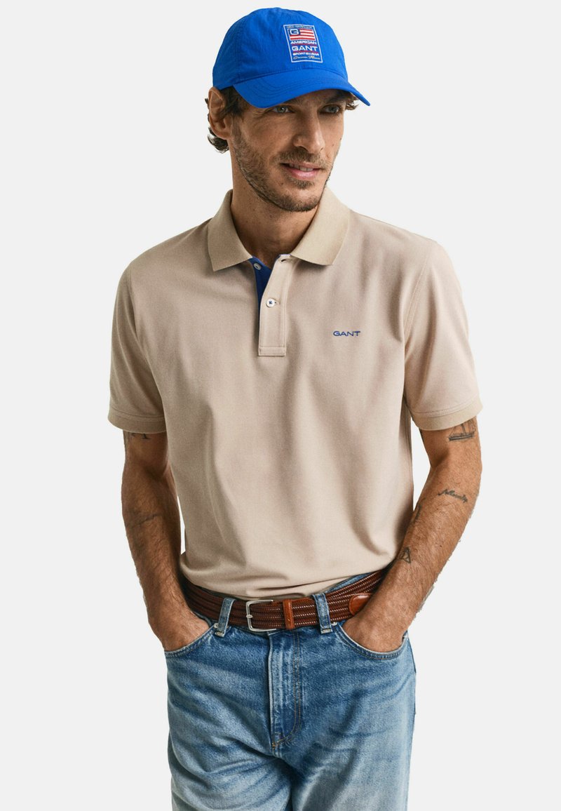 Beige polo t-skjorte med blå krage og GANT-logo. Kombinert med blå denimjeans og en blå caps med logo-patch. Brun belte.