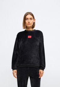 Sweatshirt em veludo preto com decote redondo, mangas longas e um emblema retangular vermelho com a palavra "HUGO" na parte da frente.