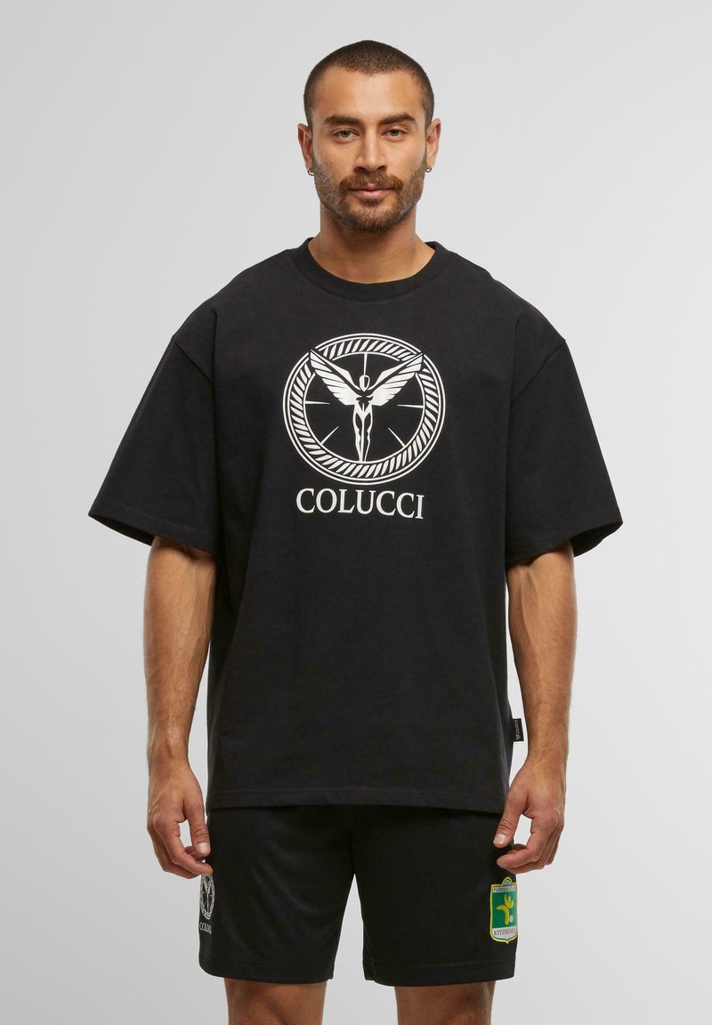 Mann trägt ein übergroßes schwarzes T-Shirt mit einer weißen geflügelten Figur und dem Text "COLUCCI", kombiniert mit schwarzen Shorts, die ein grün-gelb emblema zeigen.