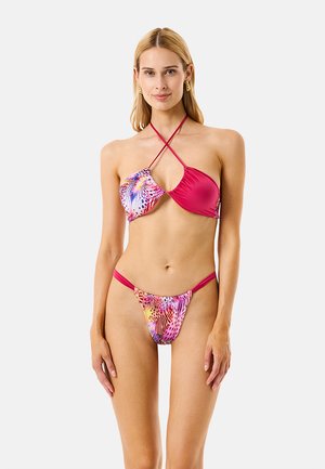 Donna che indossa un bikini colorato e asimmetrico con un top incrociato all'haltere e slip abbinati con motivo, in piedi su uno sfondo semplice.