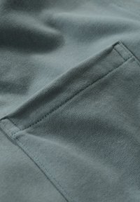 Bomullstyg i en dämpad teal-färg med en strukturerad yta, som har en broderad fickdetalj på övre vänstra sidan.