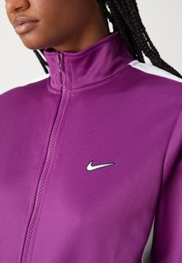 Lila jacka med dragkedja i slätt tyg, med hög krage, vita axelmarkeringar och en liten svart Nike-logotyp på bröstet.