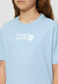 Lichtblauw katoenen t-shirt met een ronde halslijn. Bevat witte tekst: "Optimistisch is het nieuwe cool" en een kleine bloemenafbeelding. Korte mouwen.