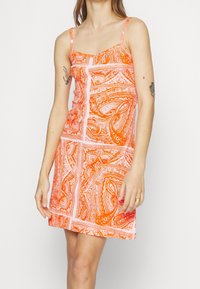 Oranssi mekko, jossa on spagettiolkaimet, kuvioitu valkoisilla pyörteillä ja viivoilla. Hieman istuva, polven ylle ulottuva pituus, pehmeä kangastunne.