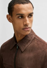 Giacca in suede marrone con colletto e zip frontale. Presenta una texture liscia e un dettaglio di cucitura minimale.