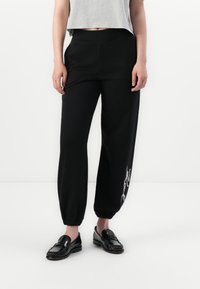 Sorte sweatpants med elastiske mansjetter, høy midje og en glatt tekstur. Har en hvit kursiv trykkdetalj på venstre ben.