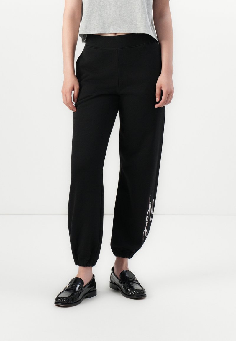 Sorte sweatpants med elastiske mansjetter, høy midje og en glatt tekstur. Har en hvit kursiv trykkdetalj på venstre ben.