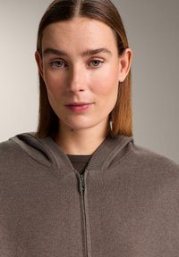 Bruine zip-up hoodie met een zachte textuur en geribbelde afwerking bij de capuchon. Daaronder is een donkerbruine top zichtbaar. Neutrale achtergrond.