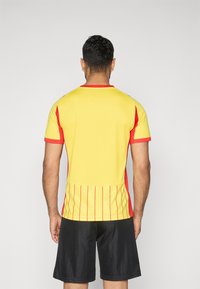 Una persona è in piedi di spalle, indossando una maglietta sportiva gialla con accenti rossi e pantaloni neri, su uno sfondo grigio uniforme.