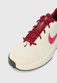 Beige atletiske sko med en tekstureret mesh-overdel, burgunderrøde snørebånd og et lyserødt Nike-logo. Gummisålen har riller for bedre greb.