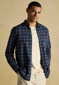 Sasietais flaneles krekls zilā, brūnā un krēmkrāsas rūtiņās. Garas piedurknes, priekšējās kabatas, valkāts atvērts virs gaiši beidzēta krekla. Strukturēta auduma.