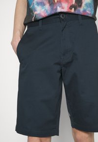 Shorts bleu marine en tissu mélangé coton, avec un devant plat, des poches latérales et une fermeture à bouton avec des coutures contrastées.