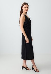Junge Frau in einem ärmellosen schwarzen Midi-Kleid und schwarzen High-Heels, die seitlich vor einem schlichten hellen Hintergrund steht.