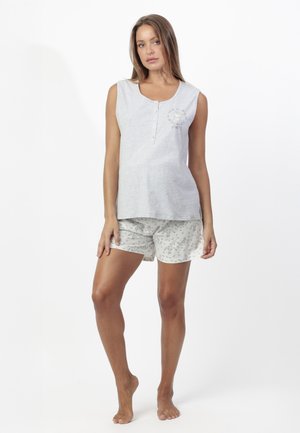 Mujer joven de pie descalza, vistiendo una camiseta sin mangas de color gris claro y pantalones cortos blancos con estampado, sobre un fondo blanco.