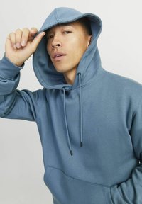 Blå hætte sweatshirt med en frontlomme, snore og en mediumvægtet, glat tekstur. Modellen justerer hætten.