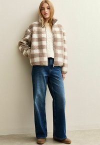 Fleece zip-up jacka i beige och vit rutmönster, kombinerad med blå vida jeans. Strukturerat material, hög krage och avslappnad passform.