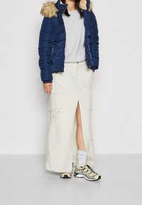 Niebieska pikowana kurtka z kapturem obszytym futerkiem, zestawiona z długą kremową spódnicą z rozcięciem z przodu i dwoma kieszeniami cargo. Całość dopełniają beżowo-czarne trampki.