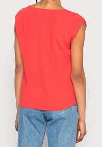 Rød kortærmet top med blonde detaljer på skulderen, glat tekstur, afslappet pasform, længere bagtil, parret med lyseblå jeans.