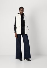 Vit pufferväst med huva, som har vertikal sömnad, zip-lock fickor och svarta detaljer, tillsammans med mörka denim flared byxor.