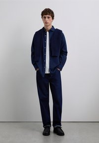 Camisa azul marino abotonada, superpuesta con un suéter de punto azul marino, camiseta blanca debajo, vaqueros oscuros y zapatos negros; estilo casual relajado.