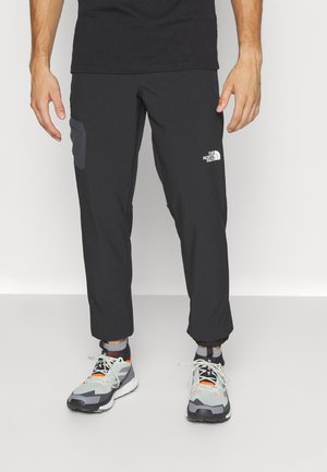 Mężczyzna w czarnych spodniach North Face z boczną kieszenią oraz szarych, białych i pomarańczowych butach sportowych stojący na szarej podłodze.