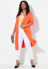 Lång orange kofta med fickor över en blommig blus, parat med vita slim fit-byxor och svarta högklackade skor, hållande en clutch i handen.