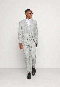 Grauer Karoanzug mit tailliertem Sakko und Hose, kombiniert mit weißem Hemd und schwarzen Schuhen. Accessoires umfassen eine schwarze Sonnenbrille.