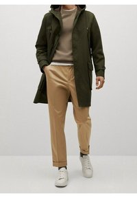 Olijfgroene, weersbestendige jas met capuchon, beige gebreide top, tan broek en witte sneakers. Bevat zakken en knoopaccenten.