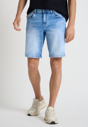 Mann, der hellblaue Denim-Shorts, schwarzes Hemd und beige Sneaker trägt, steht vor einem einfarbigen weißen Hintergrund.