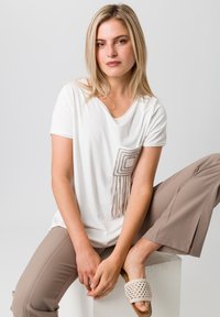 Witte shirt met korte mouwen en een decoratieve tan kleurige zak met franjes, gecombineerd met beige broek en geweven beige sandalen.