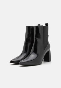 Paire de bottines noires brillantes à bouts pointus, avec des panneaux latéraux élastiques et des talons hauts épais sur fond blanc.