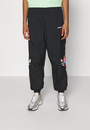 Osoba nosząca czarne spodnie dresowe Adidas z kolorowymi naszywkami z logo na kolanach i srebrno-białe sportowe buty, stojąca na szarej podłodze.