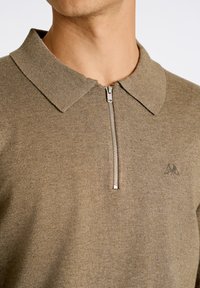 Beige zip-up pikétröja med krage och stickad struktur. Har en subtil logotyp på bröstet och detaljer i silverfärgad dragkedja.