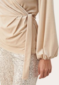 Top de terciopelo beige con diseño de envoltura y mangas abullonadas; combinado con pantalones de lentejuelas de colores claros, que presentan una textura brillante y reflectante.