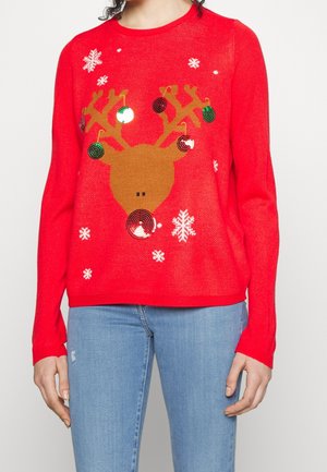 Mujer con un suéter rojo de Navidad que tiene la cabeza de un reno decorada con adornos colgantes y copos de nieve blancos.