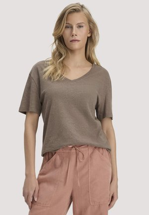 Blonde Frau mit lockerem, welligem Haar, die ein taupefarbenes V-Ausschnitt-T-Shirt mit kurzen Ärmeln und hoch taillierte, staubig-rosa Schnürhosenhosen trägt.