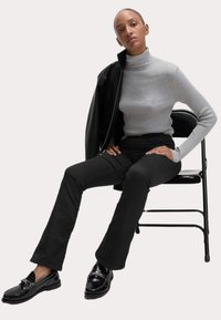 Grå ribbet turtleneck, sorte tætsiddende bukser og glinsende sorte loafers med kædedetalje; siddende på en sort sammenklappelig stol.