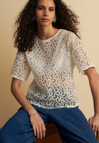 Haut en dentelle florale blanche à manches courtes, avec un design transparent et un col festonné. Associé à un jean en denim bleu.