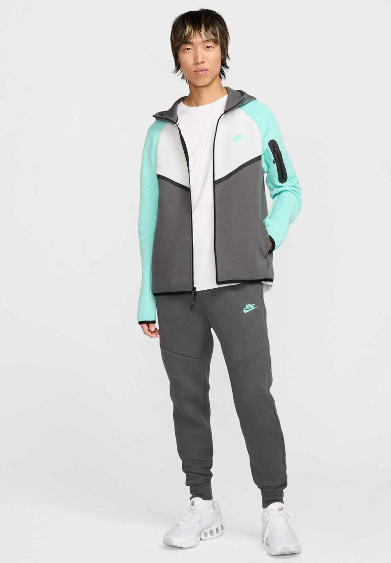 Ropa deportiva Nike que incluye una chaqueta con cremallera en color gris y menta, con un panel blanco, combinada con pantalones de chándal grises y zapatillas blancas.