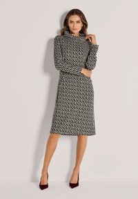 Frau in einem knielangen, langärmligen Kleid mit schwarz-weißer geometrischer Musterung, hohem Kragen und bordeauxfarbenen High Heels.